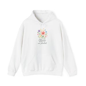 May include: White hoodie with a front pocket and drawstring hood. The front features a floral design with the text "Bloom where you are planted" in a script font. The flowers are in shades of pink, yellow, blue, and purple.