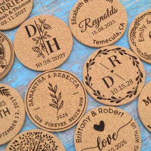 Puede incluir: Diez posavasos redondos de corcho con varios diseños y textos, incluyendo nombres, fechas y frases relacionadas con la boda. Los posavasos son de color marrón claro y tienen una textura de corcho natural.