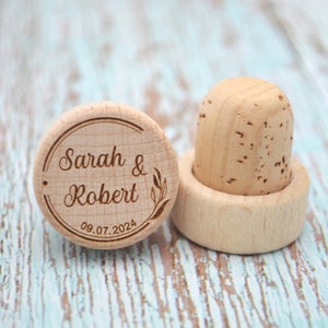 May include: Two wooden bottle stoppers, one with a cork top and the other with the inscription "Sarah & Robert 08.07.2024" in a circle.
