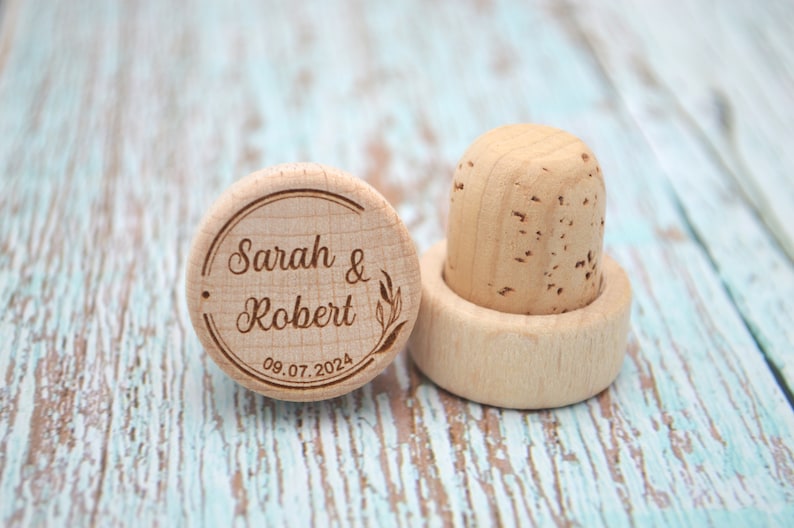 May include: Two wooden bottle stoppers, one with a cork top and the other with the inscription "Sarah & Robert 08.07.2024" in a circle.