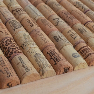 May include: Close-up of a collection of used wine corks, arranged in diagonal rows. The corks are made of natural cork material, with various printed designs and text, including brand names and logos. The corks are in shades of brown and beige.