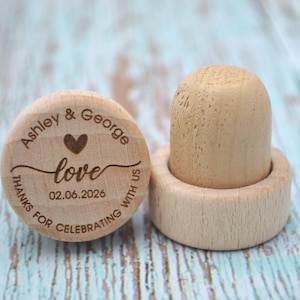 May include: Two wooden bottle stoppers with engraved text. The first stopper says "Ashley & George love 02.06.2026 Thanks for celebrating with us". The second stopper is plain.