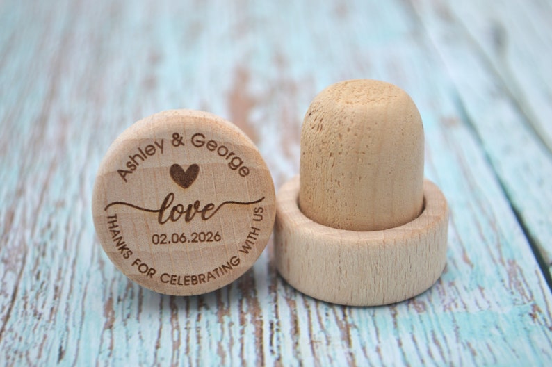 May include: Two wooden bottle stoppers with engraved text. The first stopper says "Ashley & George love 02.06.2026 Thanks for celebrating with us". The second stopper is plain.