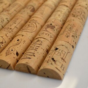 Puede incluir: Primer plano de varios tapones de corcho para vino. Los tapones son cilíndricos con extremos redondeados y están hechos de corcho marrón claro. Cada tapón tiene texto y diseños impresos, incluyendo las palabras "SHILAS" y "LOGODAJ Bulgaria".