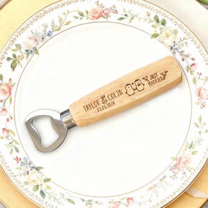 Può includere: Un apribottiglie in legno con testa in acciaio inossidabile. Il manico è inciso con "TAYLOR & COLIN 03.05.2026" e due illustrazioni di pinguini, con le parole "JUST MARRIED". L'apribottiglie è appoggiato su un piatto a fantasia floreale.