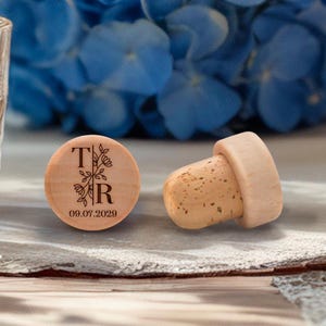 May include: Wooden bottle stopper with a cork base. The wooden top is engraved with the letters "T" and "R" with floral accents and the date "09.07.2029". The stopper is on a table with a glass and a fork.