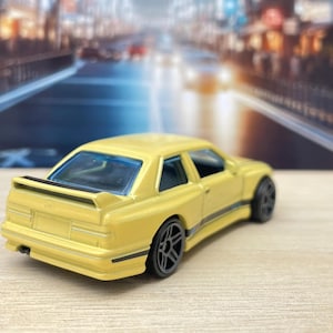 May include: A yellow die-cast toy car model of a BMW E30 M3 with black wheels and a black stripe along the side. The car is sitting on a light brown surface.