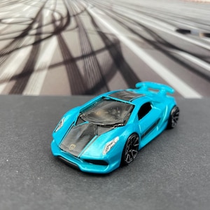 May include: A small, turquoise toy sports car with black accents on the hood and wheels. The miniature vehicle has a spoiler and is set against a blurred background of a city scene.