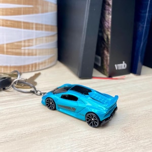May include: A blue toy car keychain with black detailing and a black stripe on the side. The car has a black windshield and black wheels. The keychain is attached to a silver metal ring.
