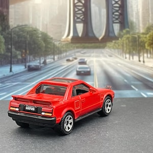 May include: A red toy car model of a Toyota MR2, with black accents and a black rear spoiler. The car is on a gray surface with a blurry background of a bridge.