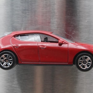 May include: A red toy car model of a Mazda 3 hatchback. The car has black wheels and a silver trim around the windows.