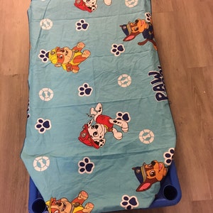 May include: A blue fitted sheet with a Paw Patrol theme. The sheet features paw prints and images of the characters Chase, Marshall, Skye, and Rubble. The sheet is made of a soft, comfortable fabric.