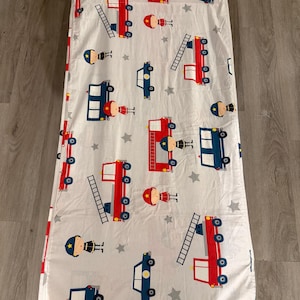 May include: A white rectangular sleeping mat cover featuring a repeating pattern of red fire trucks, blue cars, and cartoon firefighters in blue uniforms with yellow helmets, interspersed with small grey stars across the entire fabric surface.