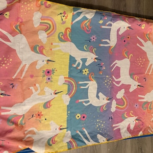 May include: A fabric sheet featuring a repeating pattern of white unicorns with multicolored manes and tails. The background consists of vertical stripes in shades of pink, yellow, and blue, decorated with small rainbows, clouds, flowers, and stars.