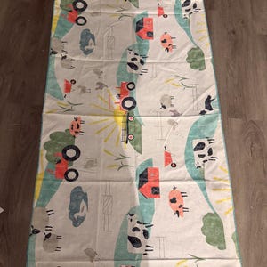 May include: A rectangular fabric mat featuring a farm-themed print on a white background. The pattern includes red barns, black and white cows, pink pigs, grey sheep, red tractors, green hills, and yellow sunbursts, all arranged in a repeating layout.