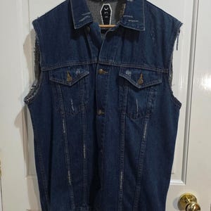 May include: A dark blue denim vest with a sleeveless design, showing distressed areas and two chest pockets. The vest has a button-up front and a frayed collar. The back of the collar has a small white graphic. It is hanging on a wooden hanger.