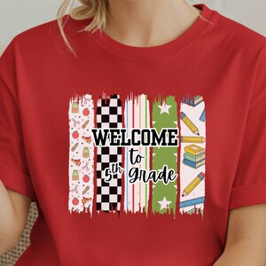 May include: A red short-sleeved t-shirt featuring a graphic print with vertical brushstroke patterns in various colors. The design includes the text "WELCOME to 5th Grade" in black lettering, surrounded by small icons of apples, pencils, and books.