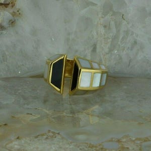 May include: A gold ring with a geometric design. The ring is made of two pieces, one black and one white, that fit together. The white piece is made of mother-of-pearl.