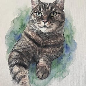 Puede incluir: Una pintura a la acuarela de un gato atigrado marr&oacute;n y gris con rayas oscuras y ojos de color verde claro. El gato aparece en posici&oacute;n sentada sobre un fondo suave y difuminado de aguadas de color verde y azul sobre papel texturizado.