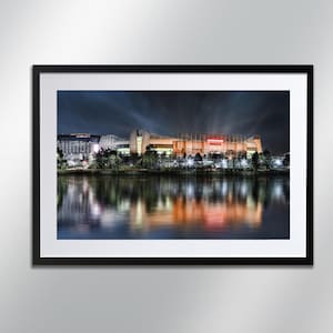 May include: A nighttime view of a large stadium with lights illuminating the building and reflecting in the water below. The stadium is surrounded by trees and buildings.