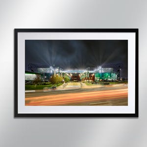 May include: A night-time view of a large stadium with lights shining down onto the pitch. The stadium is illuminated in green and white lights. Cars are driving on the road in front of the stadium, creating streaks of light.