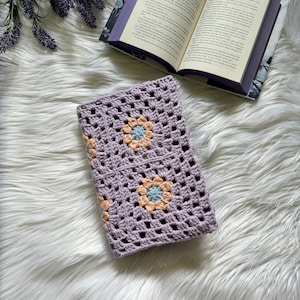 Puede incluir: Una funda de libro de ganchillo lavanda con un patrón de granny square y flores de color melocotón pastel y azul.