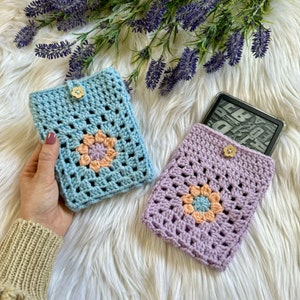 May include: Two crocheted e-reader covers with a granny square design. One cover is light blue with a peach and lavender flower in the center. The other cover is lavender with a peach and blue flower in the center.