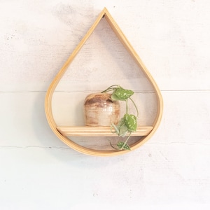 May include: A teardrop-shaped wooden wall shelf with a small ceramic pot and a trailing green plant. The shelf has a natural wood finish and is mounted on a white wooden wall. The pot is beige with brown accents.