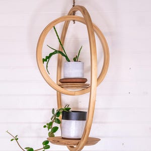 May include: A hanging wooden plant shelf with two levels, each holding a small potted plant. The shelf is made of light-colored wood and has a unique, intertwined design. The plants have green leaves and are set against a white wall.