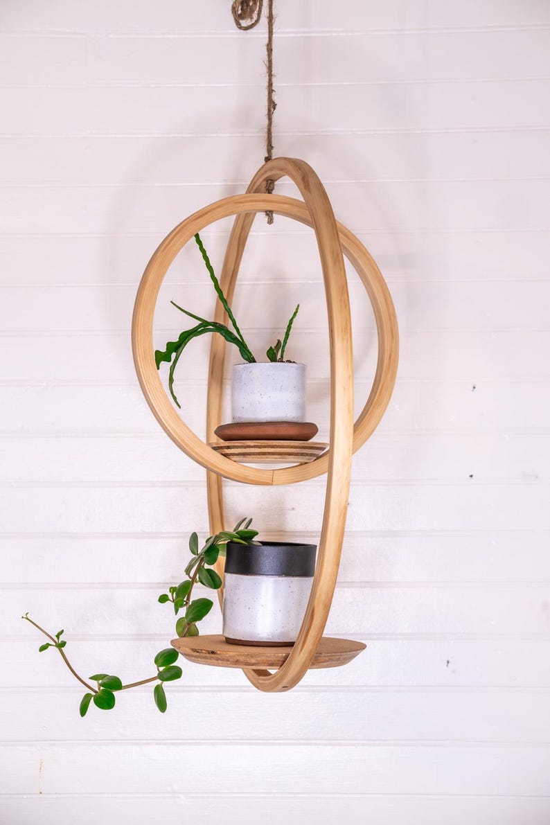 May include: A hanging wooden plant shelf with two levels, each holding a small potted plant. The shelf is made of light-colored wood and has a unique, intertwined design. The plants have green leaves and are set against a white wall.