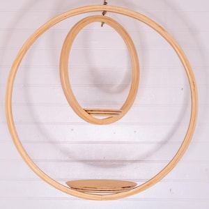 May include: A hanging wooden shelf with a circular design. The shelf features two large concentric rings, with a smaller oval shelf suspended within. A small, round shelf is positioned at the bottom. The natural wood color complements the white background.
