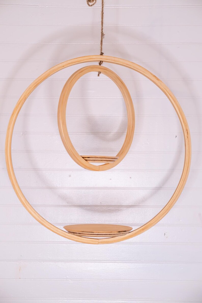May include: A hanging wooden shelf with a circular design. The shelf features two large concentric rings, with a smaller oval shelf suspended within. A small, round shelf is positioned at the bottom. The natural wood color complements the white background.