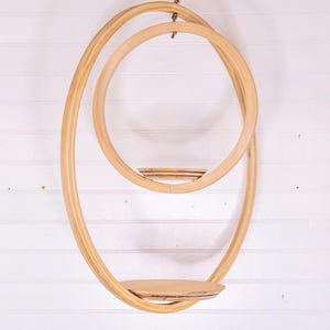 May include: A hanging wooden shelf with a unique, flowing design. The shelf features two large, intertwined oval shapes, with a small circular shelf in the middle and a larger, flat shelf at the bottom. The natural wood color complements the white background.