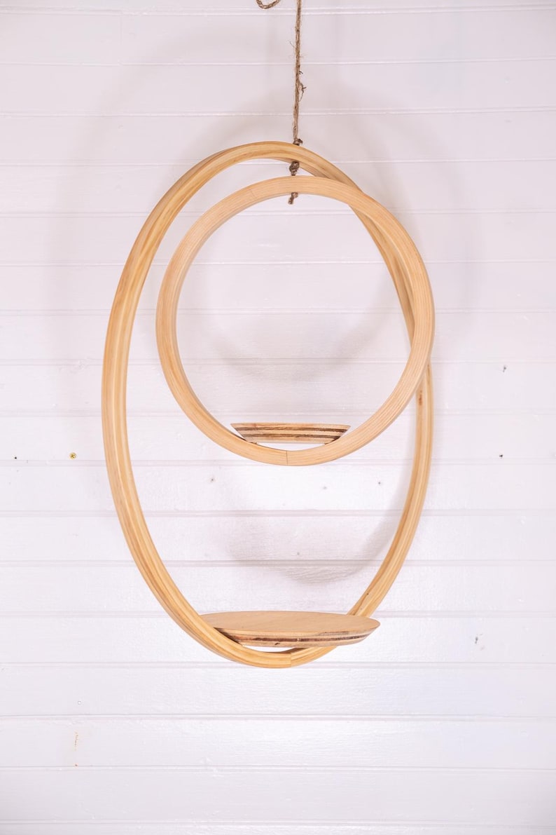 May include: A hanging wooden shelf with a unique, flowing design. The shelf features two large, intertwined oval shapes, with a small circular shelf in the middle and a larger, flat shelf at the bottom. The natural wood color complements the white background.