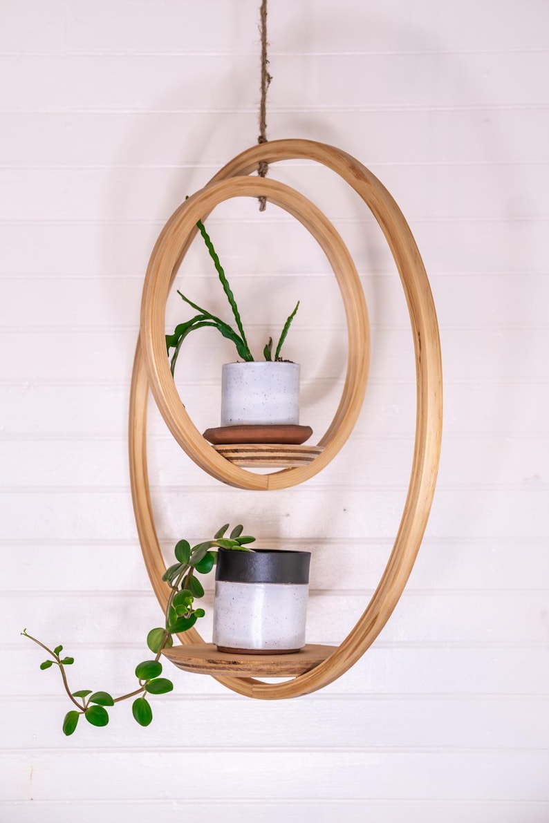 May include: A hanging wooden plant shelf with two circular tiers. Each tier holds a small potted plant. The shelf is suspended by a natural rope against a white wall. The wood has a natural finish.