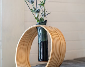 May include: A wooden vase holder with a curved design holds a dark green and black ceramic vase with blue flowers.