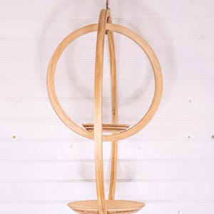 May include: A hanging wooden plant holder with a circular design. The natural wood-toned structure features a large ring, vertical supports, and two circular shelves. It is suspended by a natural fiber rope against a white background.