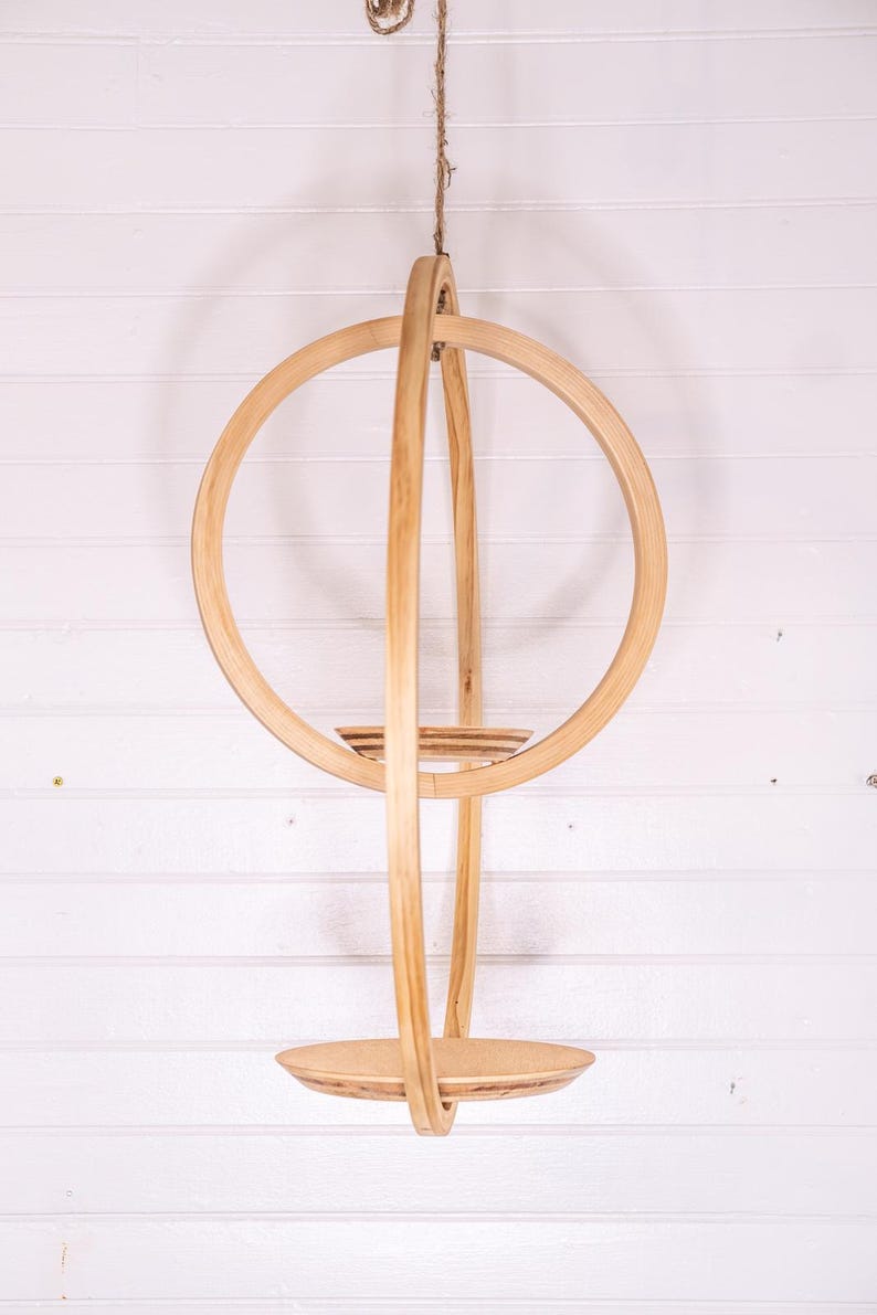 May include: A hanging wooden plant holder with a circular design. The natural wood-toned structure features a large ring, vertical supports, and two circular shelves. It is suspended by a natural fiber rope against a white background.