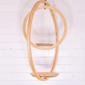 May include: A hanging wooden shelf with a circular design. The shelf is made of light-colored wood and features a round base and a series of curved, intersecting hoops. It is suspended from a natural fiber rope against a white background.