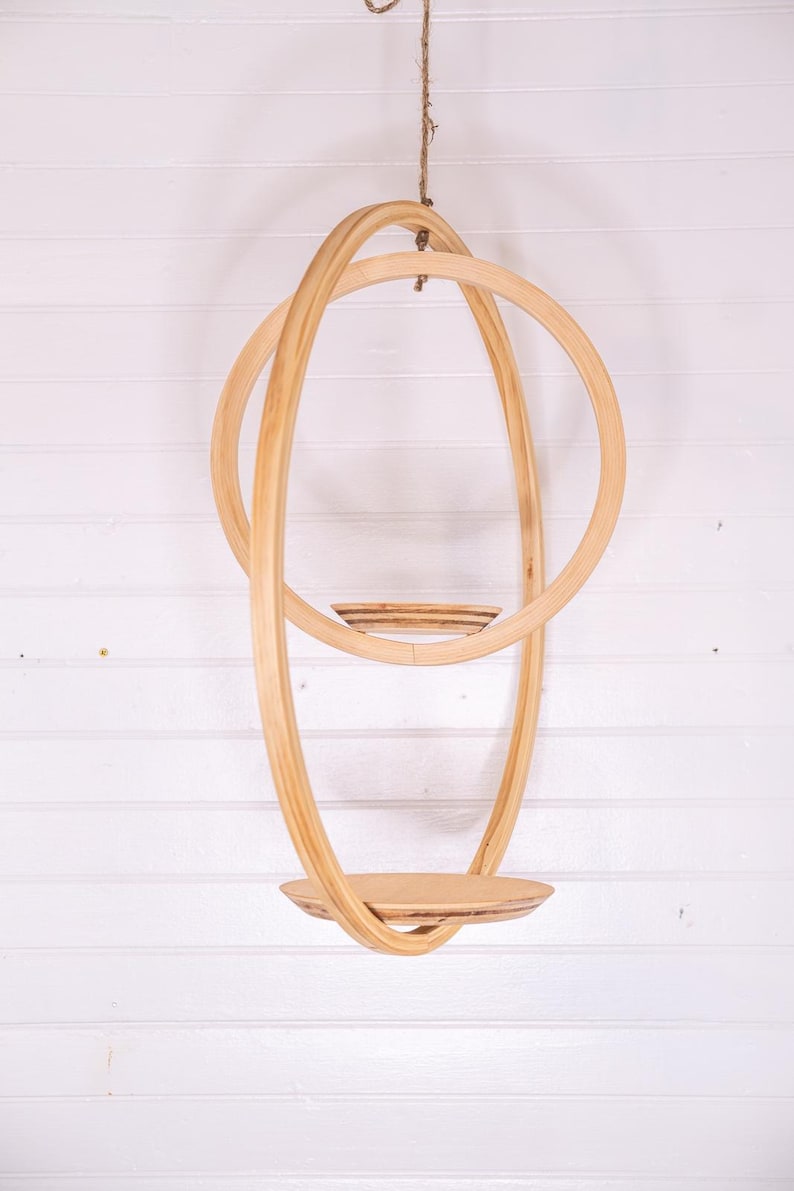 May include: A hanging wooden shelf with a circular design. The shelf is made of light-colored wood and features a round base and a series of curved, intersecting hoops. It is suspended from a natural fiber rope against a white background.
