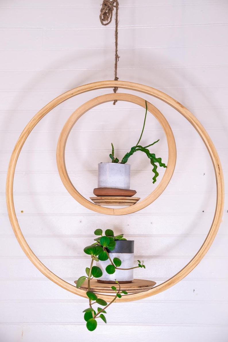 May include: A hanging wooden plant shelf with two circular shelves. Each shelf holds a small potted plant. The shelves are suspended by a natural rope against a white background. The plants have green leaves.