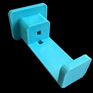 May include: A light blue 3D printed wall mount hook featuring a flat rectangular base, a protruding central block with two square-shaped openings, and a curved upward-facing end piece designed to hold items securely against a flat surface.