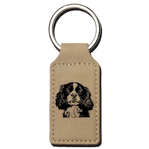May include: A tan leather keychain with a silver key ring. The keychain has a rectangular insert with a black and white silhouette of a Cavalier King Charles Spaniel.