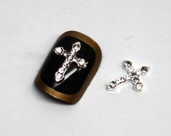 May include: Two silver cross nail charms with clear rhinestones. The cross on the left is on a black background with a gold trim.
