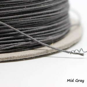 May include: A spool of mid grey elastic cord. The cord is wound tightly around the spool and a single strand of cord is hanging off the spool.