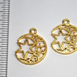May include: Two gold-tone metal charms in the shape of a crescent moon with a five-pointed star inside. The moon has a decorative filigree design.