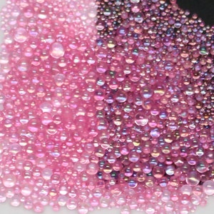 May include: A close-up image of iridescent pink and purple beads. The beads are scattered on a white surface.