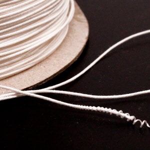 Puede incluir: Una bobina de cordón o cuerda blanca enrollada alrededor de un núcleo de cartón. El cordón está desenrollado y colgando de la bobina.