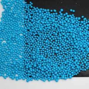 Può includere: Un'immagine ravvicinata di una grande quantità di piccole perline rotonde di colore blu turchese. Le perline sono sparse su una superficie bianca e una superficie nera.