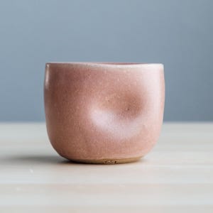 May include: A small, rounded, pink ceramic cup with a unique, dimpled design. The cup is set on a light-colored surface against a soft blue background. The cup's exterior has a smooth, matte finish.